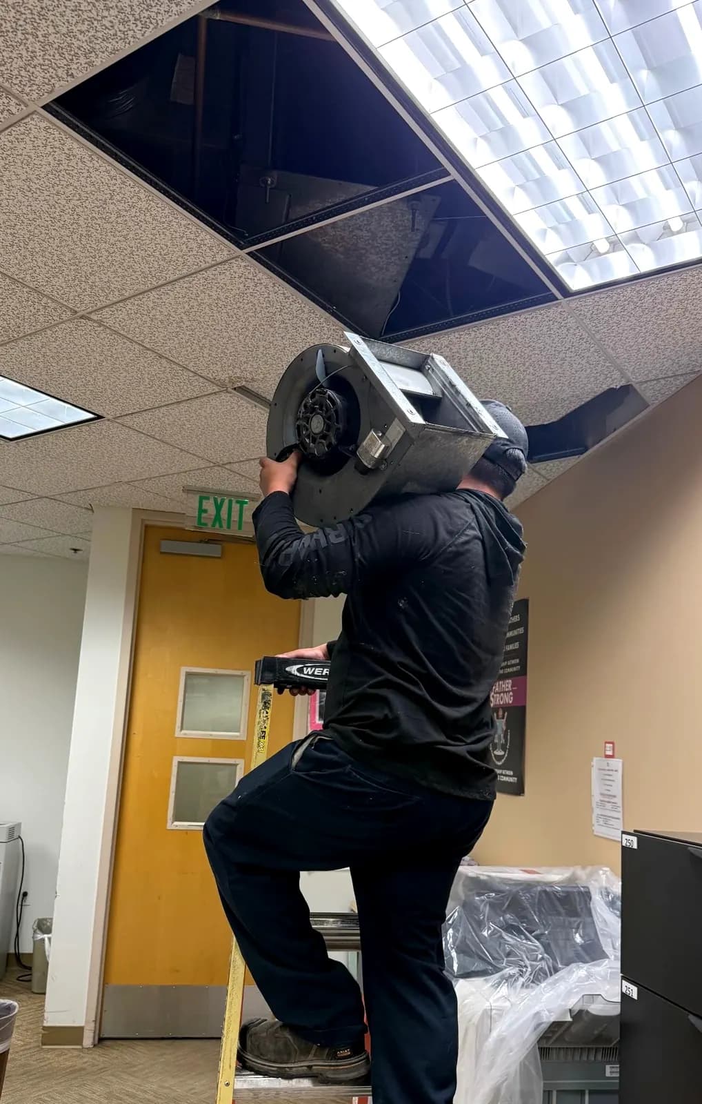 Technician carrying a blower assembly down from an office ceiling